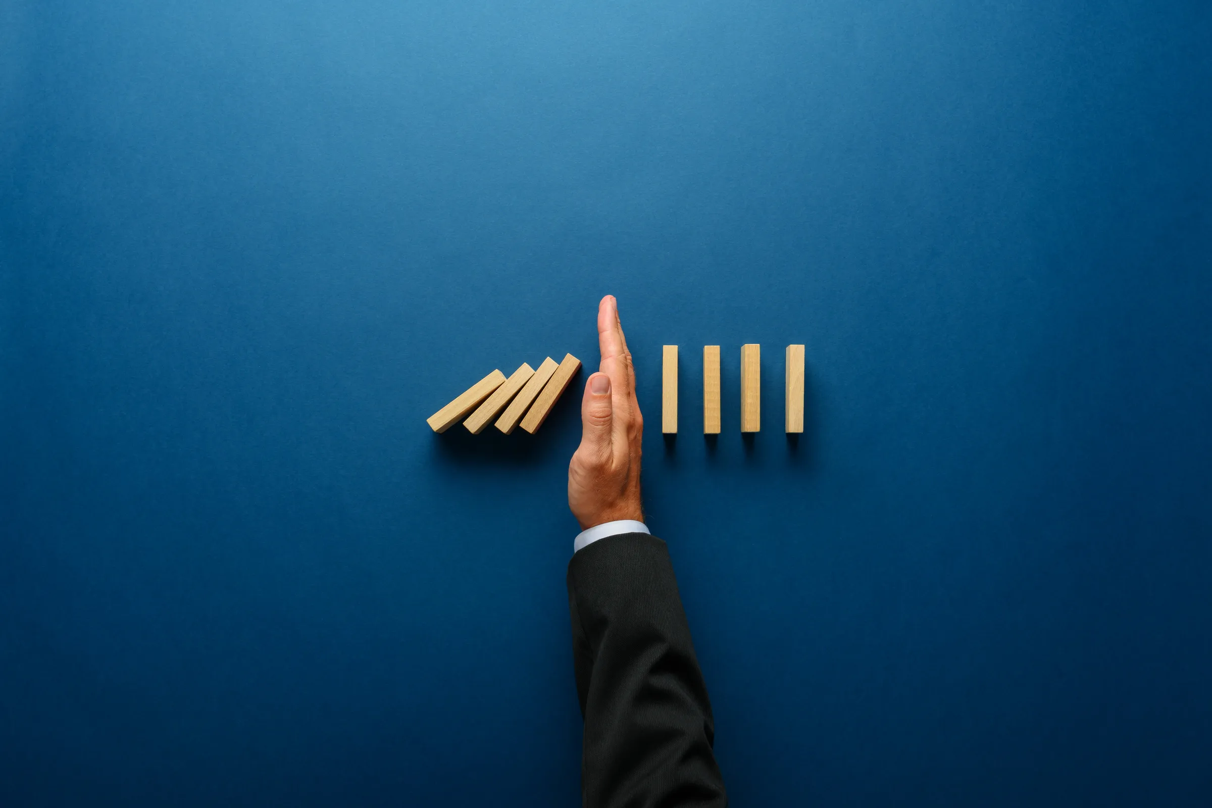 Hand in a suit sleeve stopping a row of falling wooden dominoes on a blue background