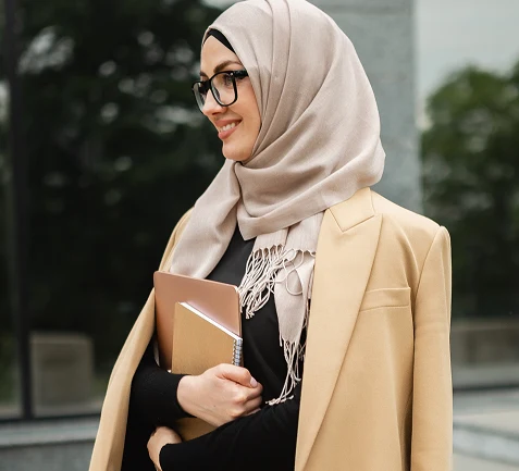Professional woman in a hijab and camel blazer holding notebooks outside an office